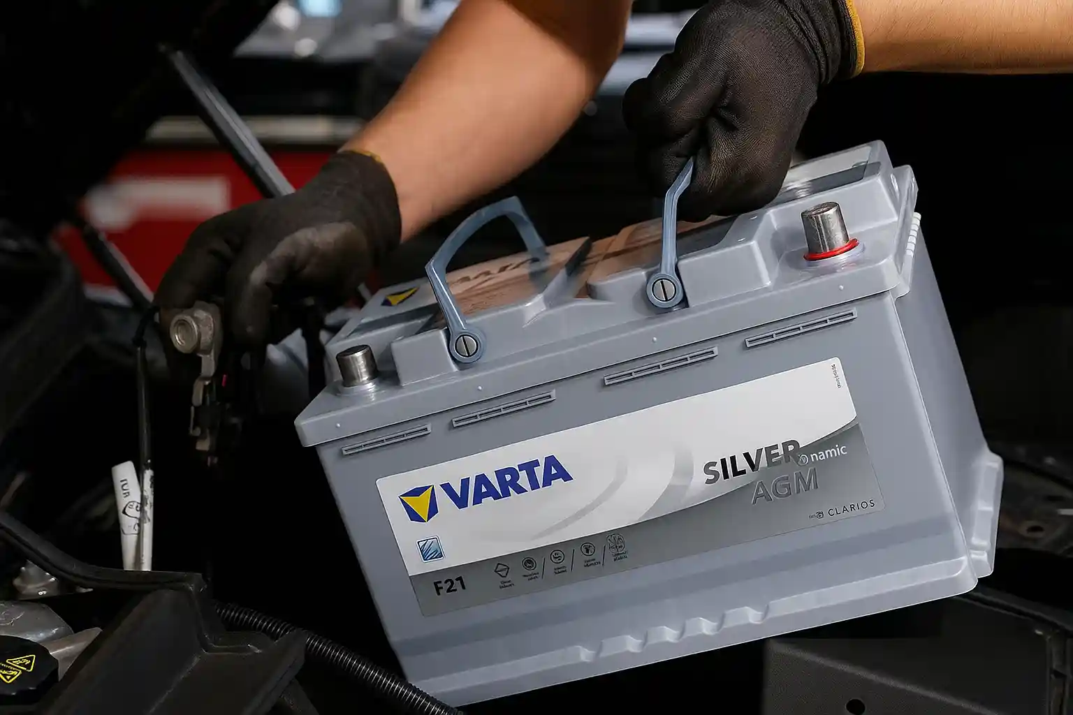 Gloved hands carrying a Varta Silver battery to an open engine bay
